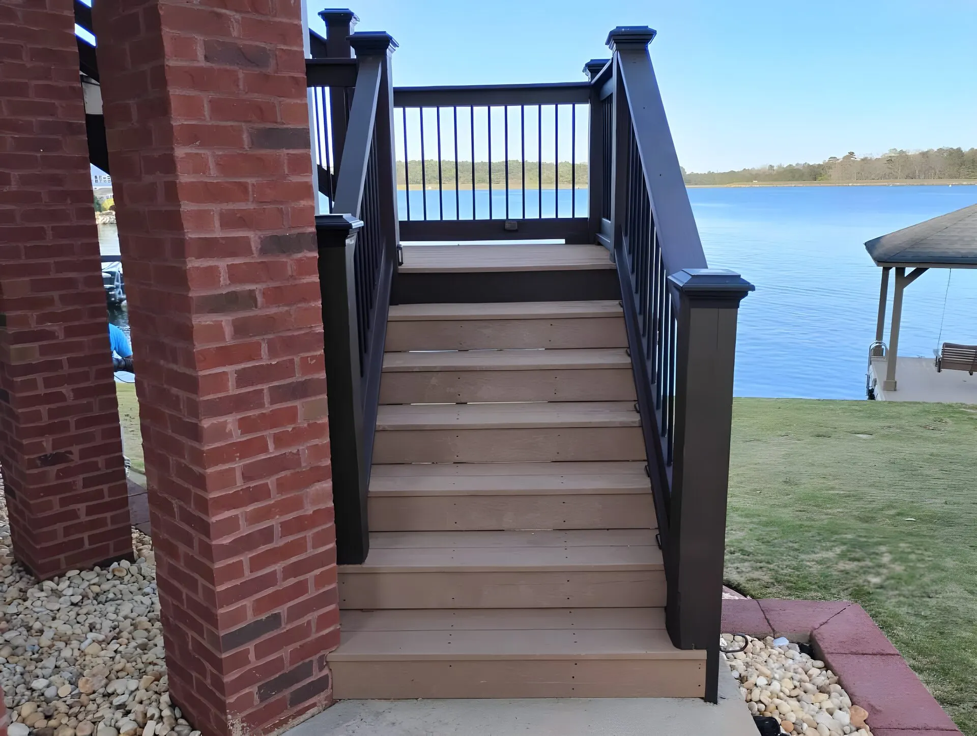 Composite deck stairs with black rails between brick columns, wide blue lake and a boathouse beyond
