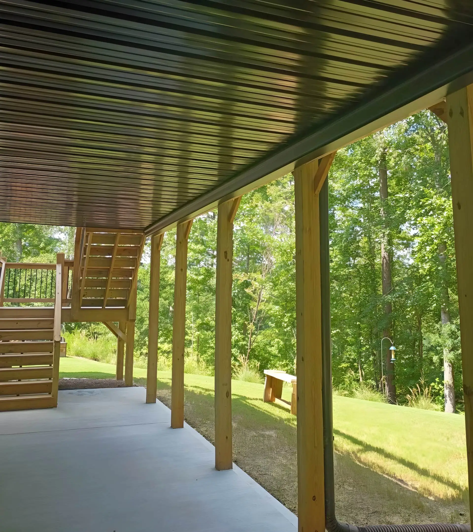 Covered patio with a corrugated metal ceiling and wood posts, stairs to an upper deck and wooded yard beyond