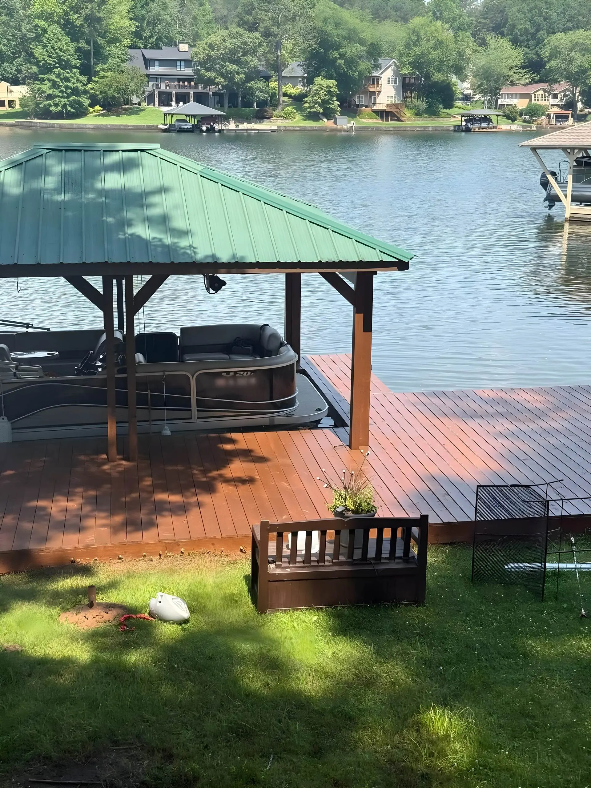 Covered boathouse with a green metal roof and richly stained dock decking, pontoon in the slip, lakefront homes across the water